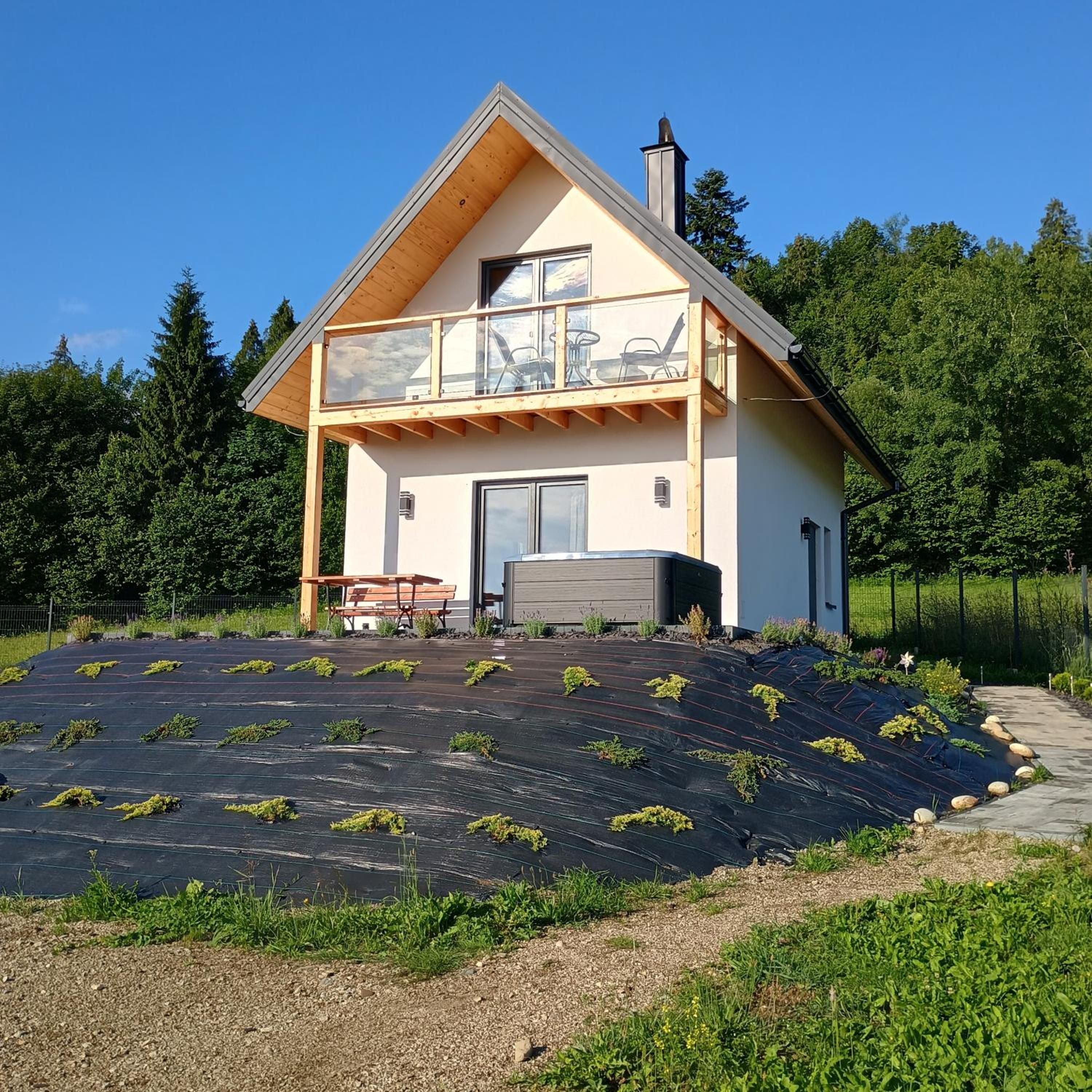 Biały Słoneczny domek z drewnianym balkonem i wanną z hydromasażem, za nim las i błękitne niebo